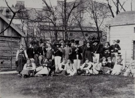 Höstfest på Kulturen någon gång på 1890-talet. Deltagarna står mellan gethuset och Mocketorpsboden med Lekloftet och Weibullska huset i bakgrunden. Alla är klädda i folkdräkt (säkerligen ur museets samlingar), sminkade med rosiga kinder, männen har käppar med bladbuketter, kvinnorna blomsterkransar. I mitt en orkester, mannen med fiol är spelmannen Ored Andersson. Stående tredje från höger ses Georg Karlin. Samtliga deltagare är män - det var studenter från Lunds universitet som grundade Kulturen vid en tid när kvinnor inte kunde studera där.