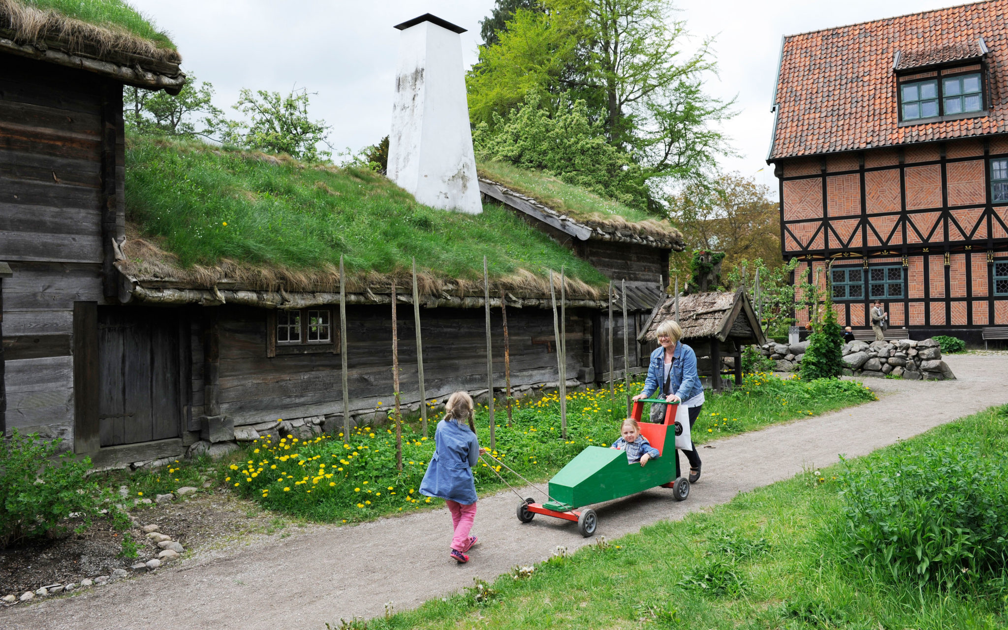 Två barn och en vuxen i friluftsmuseet, framför Blekingegården. Det ena barnet sitter i en lådbil.