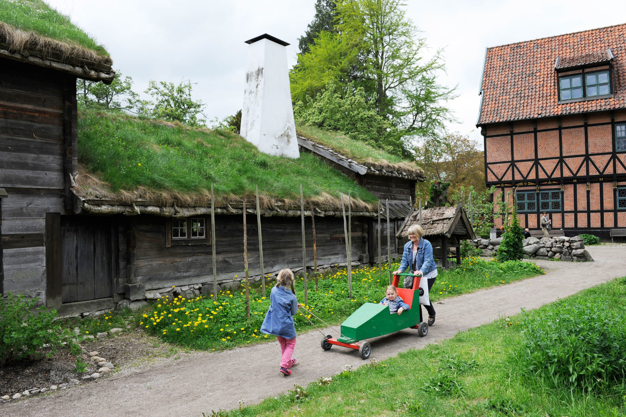 Två barn och en vuxen i friluftsmuseet, framför Blekingegården. Det ena barnet sitter i en lådbil.