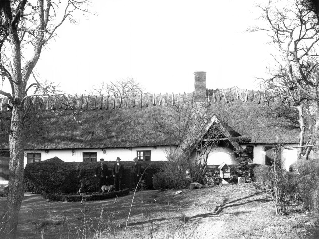 Svartvit bild av Gamlegård. Tre män i gammeldags kläder står framför korsvirkeslängan. De har en hund med sig. 