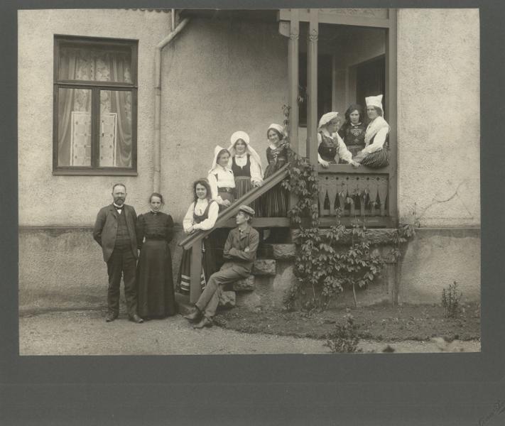 Fotografi, svartvitt, Jöns Mårtensson och hans fru Sophia framför bostaden, Villa Apla, i Lund. bredvid modern står dottern Signe, sittande sonen Torsten och näst längst till höger dottern Ingeborg. Ytterligare fem kvinnor i folkdräkt. Fotograf Lina Jonn. Daterat 1907