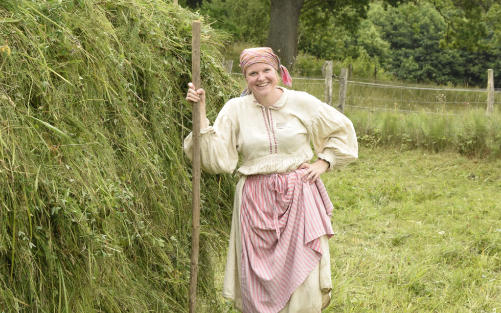 kvinna klädd som piga på 1800-talet med räfsa framför hässja med hö