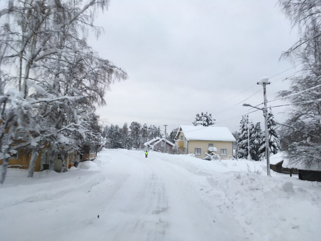 Ett snötäckt landskap med ett hus i bakgrunden och en människa som går på den snötäckta vägen. 