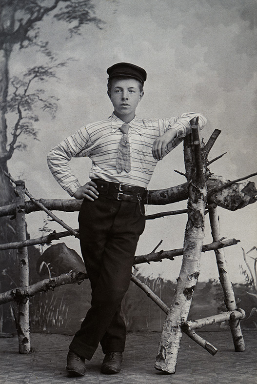 Den unge mannen är fotograferad i Umeå i slutet av 1800-talet. En del av studion har förvandlats till en utomhusscen med träd i bakgrunden och ett staket av björk. Det syns att han är stolt över sina fina kläder: skärmmössa, tvärrandig skjorta, kort slips med kravattnål, svarta långa byxor och kängor. Bältet har dubbla knäppen strax till höger syns klockkedjan. Fotograf Hanna Lundborg. Visas i utställningen "Klädd för porträtt" på Kulturen i Lund. 