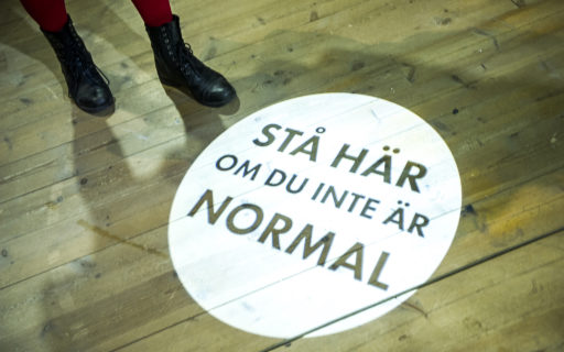 Upplyst cirkel där det står "Stå här om du inte är normal". Intill ser man någons fötter.