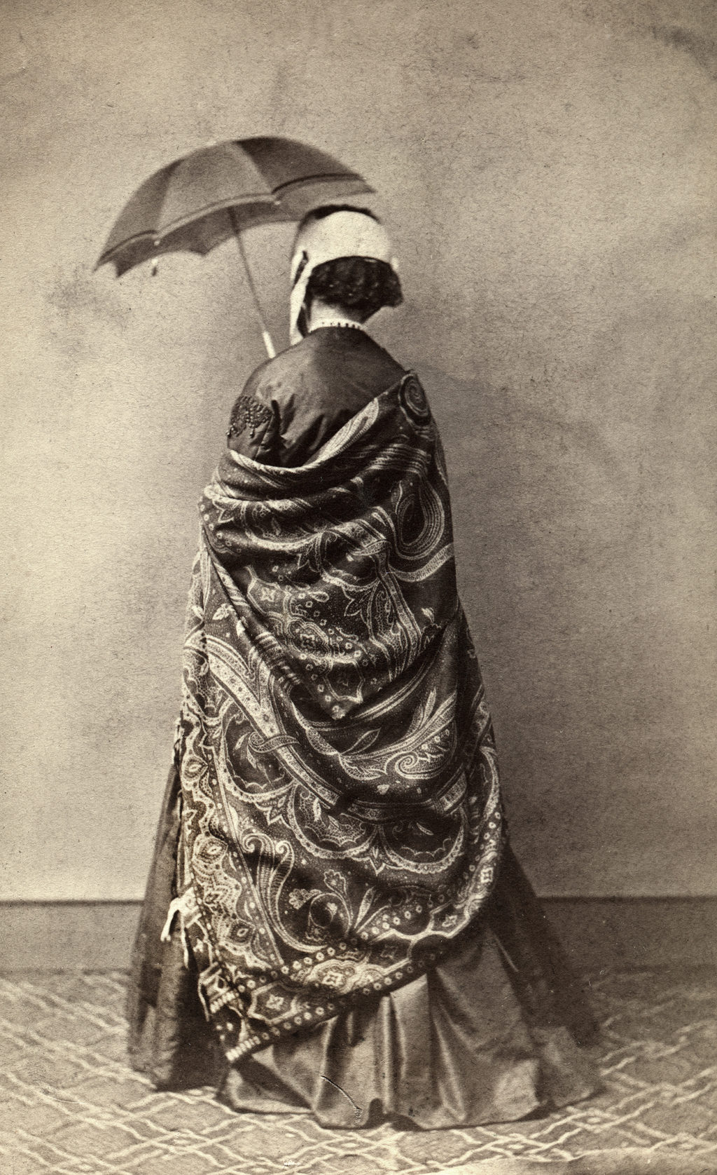 Fotografi från 1860-talet på kvinna med ryggen vänd mot betraktaren. Hon är klädd i klänning och en stor paisleysjal och håller i ett parasoll. Från utställningen Klädd för porträtt på Kulturen i Lund.