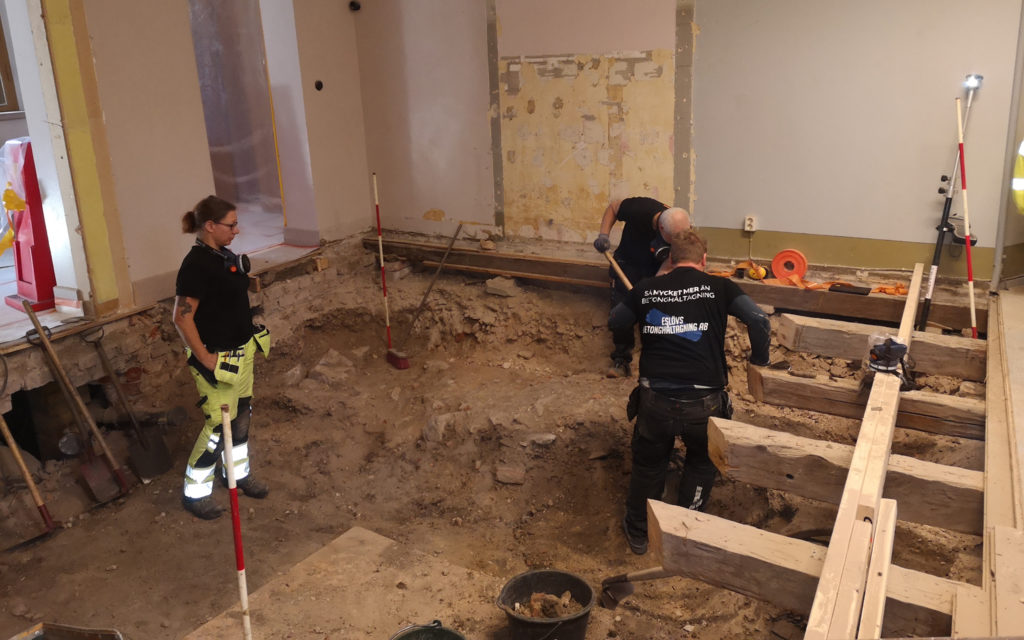 Arkeolog Linda Billström schaktningsövervakar det tunga arbetet med att hugga loss hårt sammanpressat rivningsmaterial som lagts under golvets bjälklager. Foto Kulturen. 