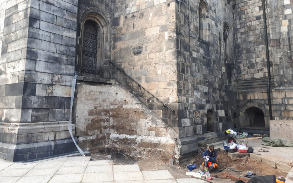 Platsen för undersökningen, vid Domkyrkans södra transept. Osteolog Helene Wilhelmson (Sydsvensk Arkeologi) undersöker graven i tegelkistan. Stenkistan påträffades senare, rakt nedanför dörren. Foto: Adam Hultberg, Kulturen. 