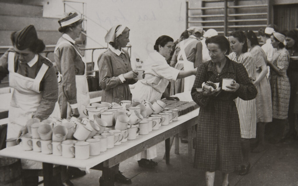 Personal i vita förkläden och hättor delar ut mat och dryck till kvinnor i 40-talsklänningar. Svartvit bild.