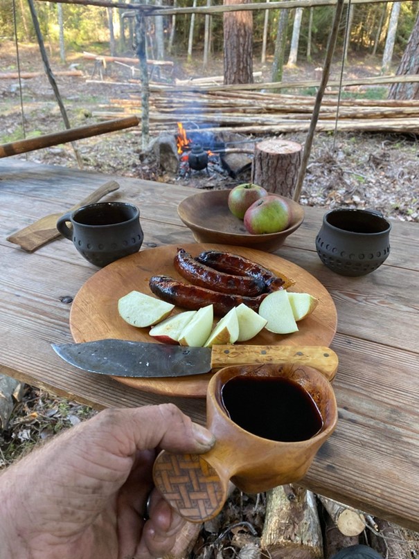 Kaffe, korv och äpplen framtdukade på en träbräda. I bakgrunden en öppen eld med kaffepanna. 