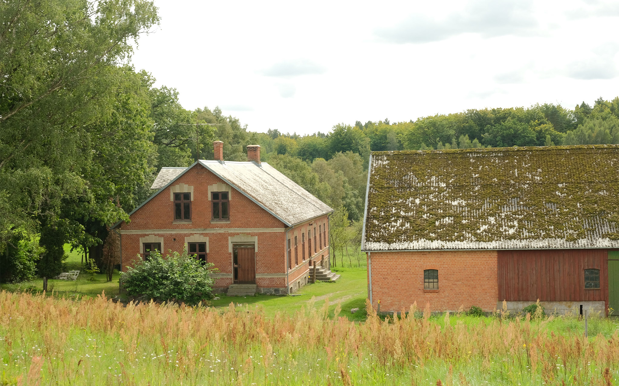 Två röda tegelbyggnder omgivna av träd, ett bostadshus och en del av en ekonomibyggnad.