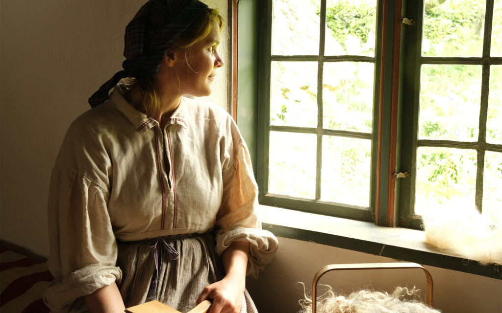 Kvinna klädd i allmogekläder tittar ut genom ett fönster.