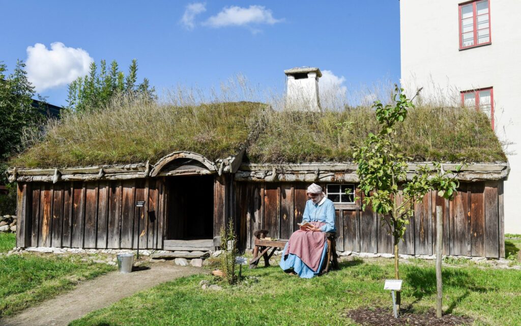 Kvinna i lång blus och lång kjol, förkläde och sjalett på huvudet sitter utanför ett litet torp.
