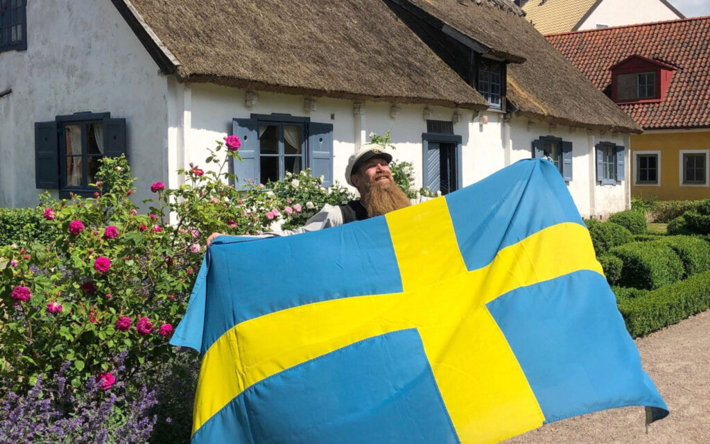 En man håller upp en stor svensk flagga. Han står framför ett kulturhistoriskt hus i friluftsmuseet på Kulturen i Lund.