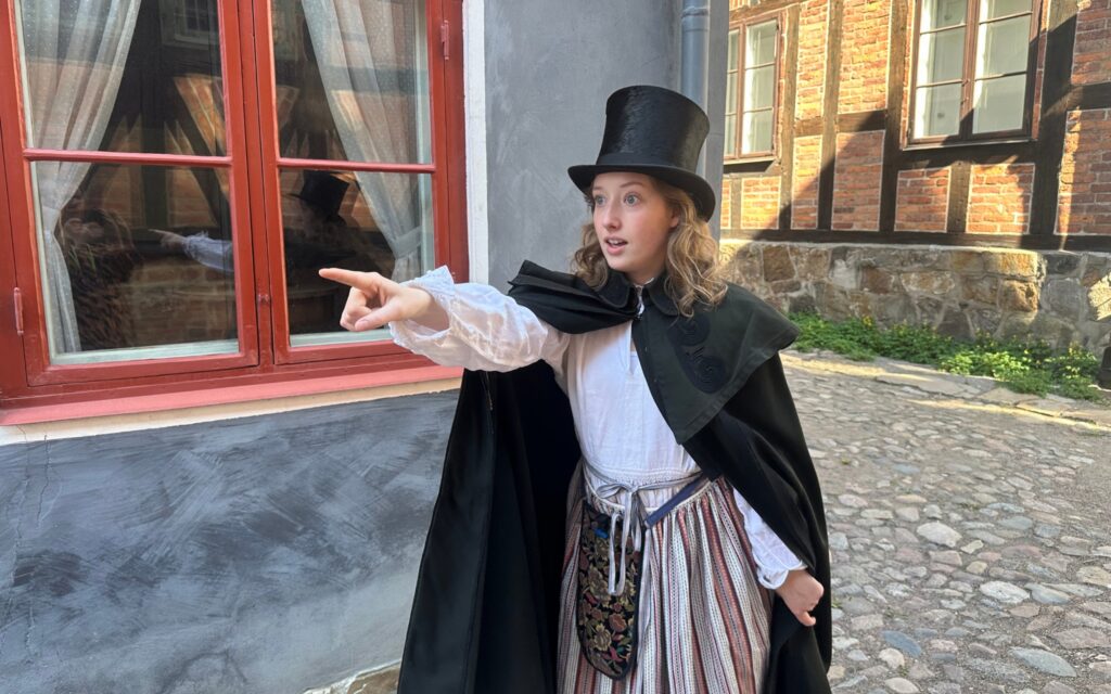 En ung kvinna i hög hatt, cape, vit blus, randigt förkläde och broderad midjeväska står framför ett gråblått hus med rödmålade fönster och pekar mot något framför sig.